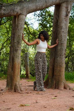Leopar desenli pantolonlu bir kadın ağacın önünde poz veriyor. Görüntü doğal, toprak hissi veriyor. Çamurda duran kadın ve ağaç topraklama hissi veriyor.