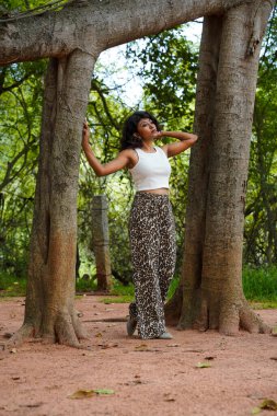 Beyaz atlet ve leopar desenli pantolon giyen bir kadın iki ağacın önünde poz veriyor.