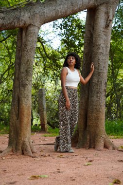 Ağacın önünde beyaz bir atlet ve leopar desenli pantolon giymiş bir kadın duruyor.