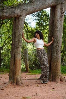 Bir kadın, bir ağacın önünde kollarıyla poz veriyor. O, beyaz bir atlet ve kahverengi pantolon giyiyor. Bu görüntünün doğal ve huzurlu bir havası var.