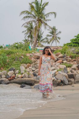 Sahilde renkli bir elbise giymiş bir kadın yürüyor. Kumsal kayalık ve gökyüzü bulutlu.