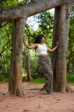 Bir kadın bir ağacın önünde poz veriyor, leopar desenli pantolon ve beyaz bir bluz giyiyor.
