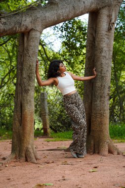 Bir kadın iki ağacın önünde poz veriyor. Beyaz bluz ve siyah pantolon giyiyor.
