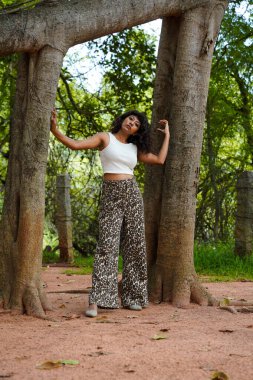 Bir kadın, bir ağacın önünde leopar desenli pantolon ve beyaz bir bluzla poz veriyor.