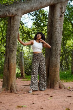 Bir kadın bir ağacın önünde kollarını kavuşturarak poz veriyor. Beyaz bir atlet ve leopar desenli pantolon giyiyor. Görüntünün doğal ve rahat bir havası var.