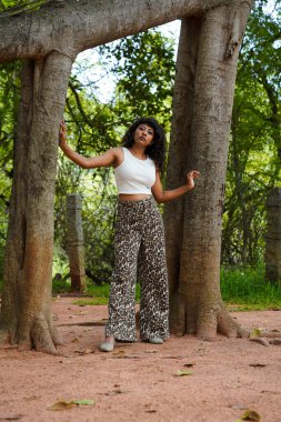 Beyaz atlet ve leopar desenli pantolon giyen bir kadın ağacın önünde poz veriyor.