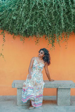 Bankta oturan bir kadın desenli beyaz bir elbise giyiyor. Yüzünde bir gülümsemeyle kameraya bakıyor.