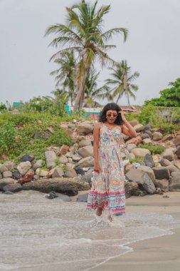 Sahilde renkli bir elbise giymiş bir kadın yürüyor. Kumsal kayalık ve gökyüzü bulutlu.