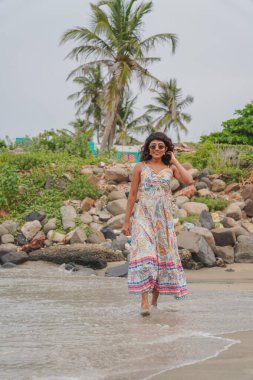 Sahilde renkli bir elbise giymiş bir kadın yürüyor. Plaj kayalar ve palmiye ağaçları ile çevrilidir. Kadın sahilde eğleniyor.