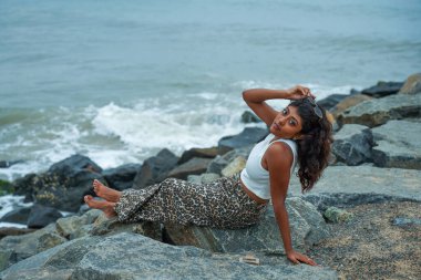 Bir kadın okyanus kıyısındaki bir kayanın üzerinde oturuyor. Leopar desenli bir etek ve beyaz bir atlet giyiyor.