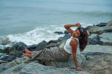 Okyanus kenarındaki bir kayanın üzerinde oturan bir kadın leopar desenli bir etek ve beyaz bir atlet giyiyor. Güneş gözlüğü takıyor ve manzaranın tadını çıkarıyor.