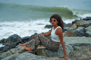 Bir kadın okyanus kıyısındaki bir kayanın üzerinde oturuyor. Leopar desenli pantolon ve beyaz bir atlet giyiyor. Kadın okyanus manzarasının keyfini çıkarırken, görüntü sakin ve huzurlu bir ruh haline sahip.