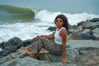 Bir kadın okyanus kıyısındaki bir kayanın üzerinde oturuyor. Leopar desenli pantolon ve beyaz bir atlet giyiyor. Kadın okyanus manzarasının keyfini çıkarırken, görüntü sakin ve huzurlu bir ruh haline sahip.