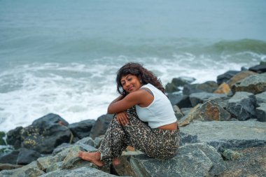 Bir kadın okyanus kıyısındaki bir kayanın üzerinde oturuyor, dalgaları seyrediyor. Leopar desenli pantolon ve beyaz bir gömlek giyiyor. Manzara sakin ve huzurlu, kadın manzaranın tadını çıkarıyor gibi.