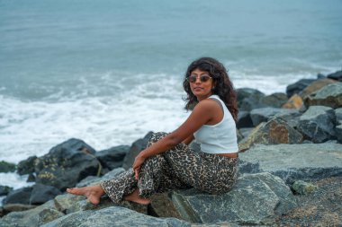 Bir kadın okyanus kıyısındaki bir kayanın üzerinde oturuyor. Leopar desenli pantolon ve beyaz bir atlet giyiyor. Sahne sakin ve huzurlu. Arka planda dalgaların çarpma sesi var.