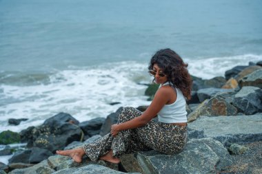Bir kadın okyanus kıyısındaki bir kayanın üzerinde oturuyor. Leopar desenli pantolon ve beyaz bir atlet giyiyor. Sahne sakin ve huzurlu. Arka planda dalgaların çarpma sesi var.