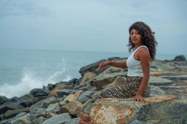 Bir kadın okyanusun kenarındaki bir kayanın üzerinde oturur ve denize bakar. Sahne sakin ve huzurlu, kadının rahat duruşu ve arka plandaki nazik dalgalarla.