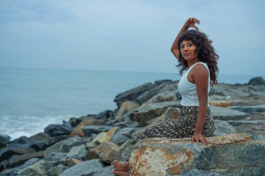 Kıvırcık saçlı bir kadın okyanusun yakınındaki bir kayanın üzerinde oturuyor. O, beyaz bir gömlek ve siyah pantolon giyiyor. Görüntü sakin ve dingin bir ruh haline sahip. Kadının duruşu ve arka planda okyanus var.