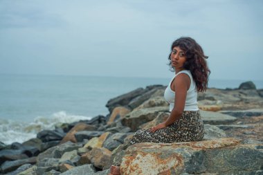 Okyanus kıyısındaki bir kayanın üzerinde oturan bir kadın. Beyaz bir atlet ve leopar desenli bir etek giyiyor. Gökyüzü bulutlu ve su sakin.