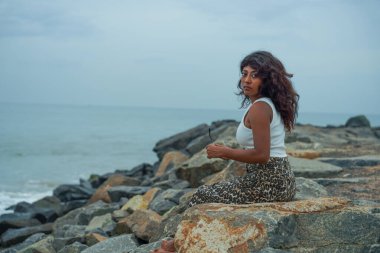 Bir kadın elinde güneş gözlüğü olan bir kayanın üzerinde oturuyor. Sahne sakin ve huzurlu, kadın suyun ve kayaların manzarasının tadını çıkarıyor.