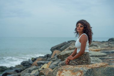 Bir kadın okyanus kıyısındaki bir kayanın üzerinde oturuyor. Beyaz bir gömlek ve leopar desenli bir etek giyiyor. Gökyüzü bulutlu ve su sakin.