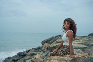 Kıvırcık saçlı bir kadın okyanusun yakınındaki bir kayanın üzerinde oturuyor. Beyaz bir atlet ve leopar desenli pantolon giyiyor. Sahne sakin ve sakin. Arka planda okyanus var.