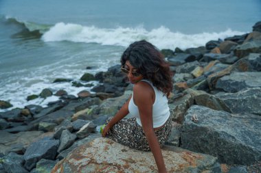 Bir kadın okyanus kıyısındaki bir kayanın üzerinde oturuyor. Beyaz bir atlet ve leopar desenli pantolon giyiyor.