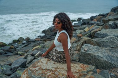 Bir kadın okyanus kıyısındaki bir kayanın üzerinde oturuyor. Beyaz bir atlet ve etek giyiyor. Sahne sakin ve huzurlu. Arka planda dalgaların çarpma sesi var.