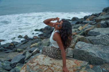 Okyanus kıyısındaki bir kayanın üzerinde güneş gözlüğü takan bir kadın oturuyor. Beyaz bir atlet ve leopar desenli pantolon giyiyor.