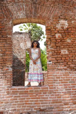Renkli bir elbise giymiş, tuğla bir duvarın önünde duran bir kadın. Tuğla duvar ve kadın elbisesi ona nostalji hissi veriyor.