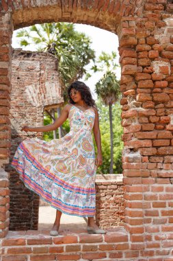Uzun elbiseli bir kadın tuğla bir duvarın önünde duruyor. Elbise beyaz ve çiçek desenli. Kadın gözlük takıyor ve fotoğraf için poz veriyor. Sahne rahat ve rahat.