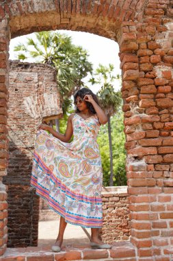 Uzun elbiseli bir kadın tuğla duvarın önünde poz veriyor. Elbise beyaz ve desenli.