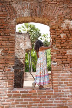 Bir kadın pencereli tuğla bir duvarın önünde duruyor. O bir elbise giyiyor ve pencereden dışarı bakıyor.