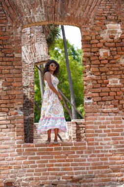 Beyaz elbiseli bir kadın tuğla bir duvarın önünde duruyor. Tuğla duvar eski ve kırsal bir hissi var. Kadın beyaz bir elbise giyiyor ve kameraya poz veriyor. Görüntünün bir mahsulü var.