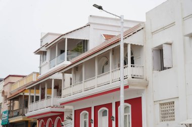 Beyaz balkonu ve beyaz sokak lambası olan uzun kırmızı bir bina. Bina diğer binalar ve caddelerle çevrili.