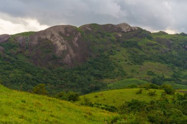 Arkasında bulutlu bir gökyüzü olan dağ sırası. Dağlar yeşil ağaçlarla çevrili.