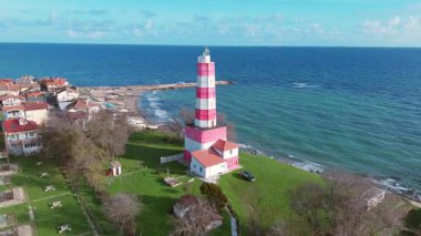 Kamera ikonik deniz feneri etrafında dönüyor, kırmızı çizgili beyaz, kayalık tepenin üzerine tünemiş.