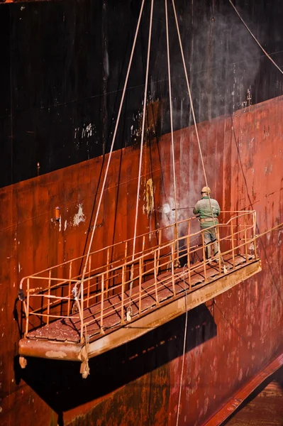 Shipyard worker power washing a ship on dry dock.