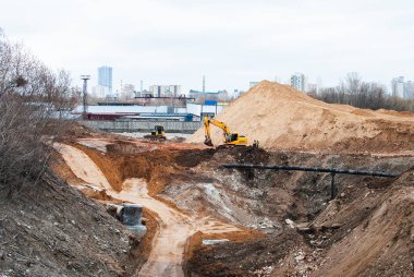 Şehir manzarası ve uzaktaki binalara karşı çukurda çalışan sarı bir kazıcı var.