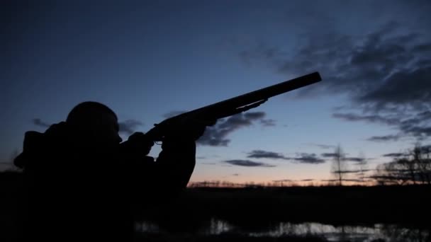 Силует мисливця вночі з пістолетом. Силует мисливця з пістолетом на фоні заходу сонця. Чоловік з пістолетом на тлі вечірнього неба і річки на полюванні — стокове відео