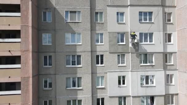 Ένας βιομηχανικός ορειβάτης αιωρείται σε σχοινιά και βάφει τον τοίχο ενός κτιρίου με ρολό. Ένας άνδρας εργαζόμενος εκτελεί εργασίες για τη μόνωση της πρόσοψης ενός πολυώροφου κτιρίου. Θέα από το drone. — Αρχείο Βίντεο