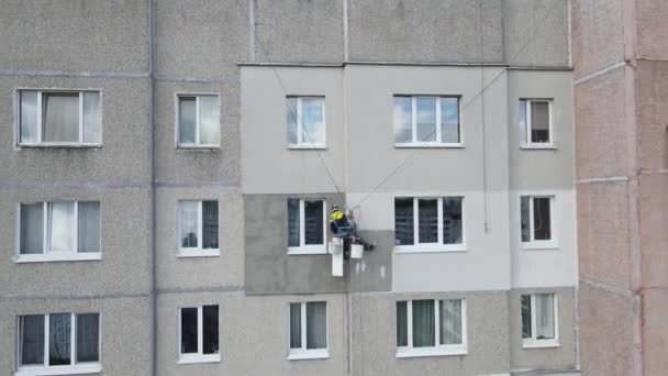 An industrial climber suspended on ropes and paints the wall of a building with a roller. A male worker performs work on the insulation of the facade of a multi-storey building. View from the drone. — Stock Video