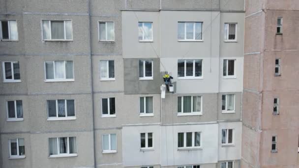 Przemysłowy alpinista zawieszony na linach maluje ścianę budynku wałkiem. Samiec wykonuje prace przy izolacji elewacji budynku wielokondygnacyjnego. Widok z drona. — Wideo stockowe