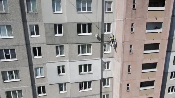 An industrial climber suspended on ropes and paints the wall of a building with a roller. A male worker performs work on the insulation of the facade of a multi-storey building. View from the drone. — Stock Video