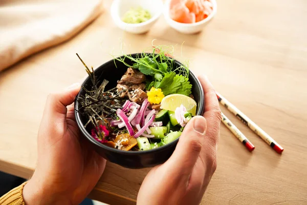 A man holds a Poke salad with beef in a bowl in his hands. Ingredients Beef, Nameko mushrooms, cherry tomatoes, rice, cucumber, red onion, sesame seeds, cilantro, lime. Asian salad concept.
