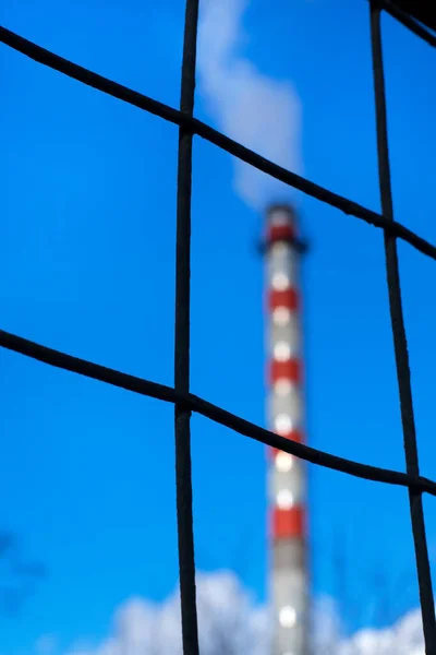 Grid metal fence and factory chimney releasing steam against the blue sky. Industrial waste