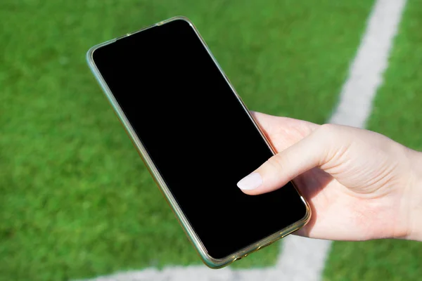 Smartphone with black screen in woman hand against the soccer field. Using mobile apps for workouts and at sports events. Copy space