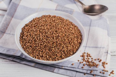 Beyaz ahşap arka planda bir kase kuru çiğ karabuğday inlemesi. Karabuğday lapası pişirme kavramı.