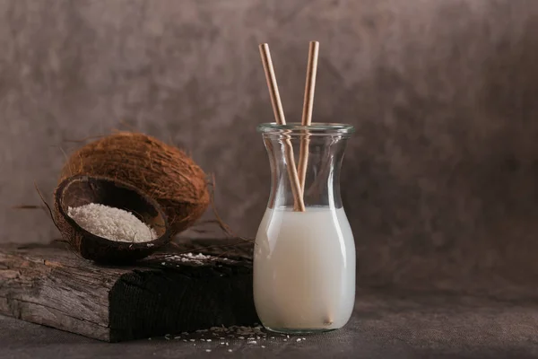 Бутылка кокосового веганского молока с соломинками, цельным кокосом и хлопьями на тёмном фоне. Понятие здорового образа жизни. — стоковое фото