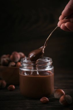 Ev yapımı çikolatalı fındıklı süt. Koyu ahşap arka planda cam kavanoza yayılmış. Kadının eli kaşığı tutuyor.